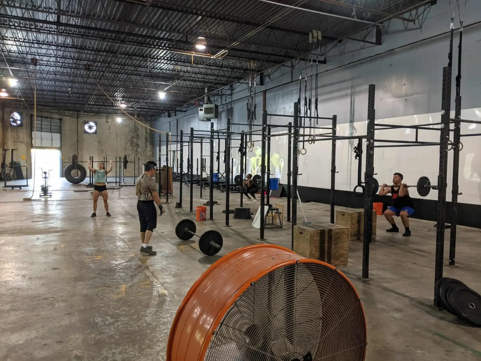Wide view of the Fit 901 training floor: bare concrete with old lifting lines, exposed steel deck ceiling, squat rigs along the right wall, a large orange floor fan in the foreground, and four members training.