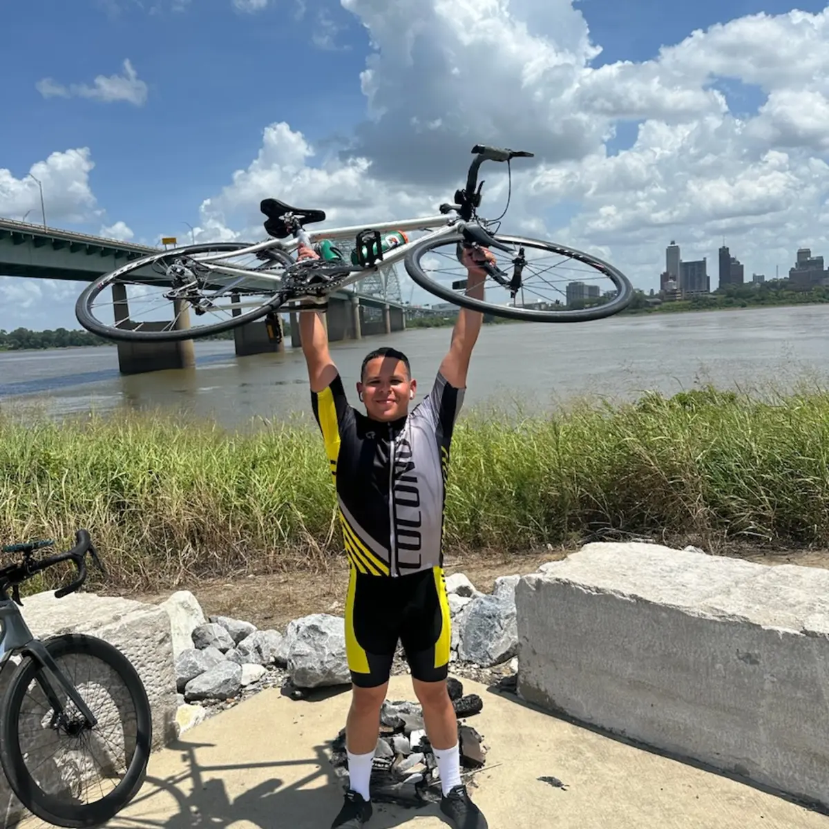 Alex holding his road bicycle above his head on the Memphis riverfront, the Hernando de Soto Bridge and the downtown skyline behind him.